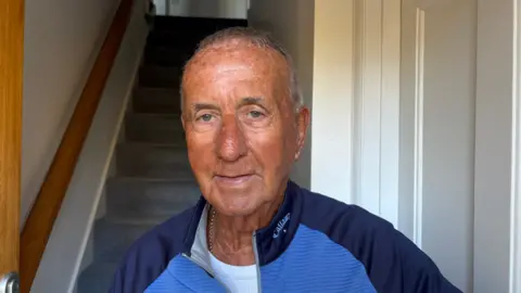 Martin Heath/BBC Derek Stokes with short white hair, smiling at the camera and wearing a thin jacket in two shades of blue. He is standing just inside his house with grey carpeted stairs behind him.