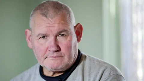 Laurence Cawley/BBC Jose looks sternly toward the camera. The wall behind him is pistachio green. He is wearing a black tee shirt and a grey sweater top with "Northfield" written on it