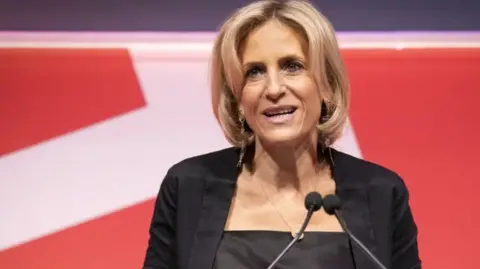 A blonde-haired woman in a dark jacket by a microphone