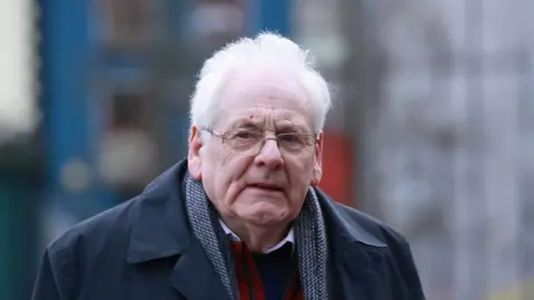 PA Michael has white hair and silver glasses. He is wearing a navy coat, navy woolen scarf and white shirt beneath a navy jumper. He is looking to the side of the camera. 