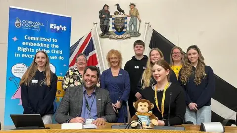 Councillor Leigh Frost sitting at a table with a group of people around him. There is a blue sign with the words 'Cornwall Council' in the top left corner. 