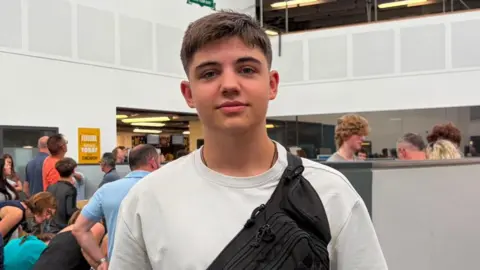 Yaroslav poses for the picture in a gymnasium  with students behind him. he is in a white shirt, and a black bag slug across his shoulder. He has brown hair 