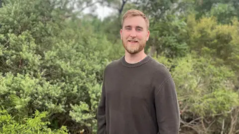 Handout Ed stands outdoors in a natural setting, wearing a dark knitted jumper and jeans. Dense green foliage surrounds him, and the ground is covered with leaves and plants. He has his hands in his pockets while positioned in front of tall shrubs and trees.