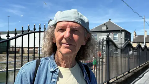 Colum Sands, a man with shoulder-length, curly grey hair, stands at the railings outside Warrenpoint Port, looking directly at the camera on a sunny day. He is wearing a blue denim shirt over a pale yellow t-shirt and a denim cap. He is carrying a black bag with a strap over each shoulder. Warrenpoint Harbour Authority's HQ - a grey two-story building - is in the background.
