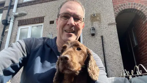 Handout A man in a blue and navy fleece takes a selfie with his dog. The man is wearing glasses and sat in front of his pebbledash house.