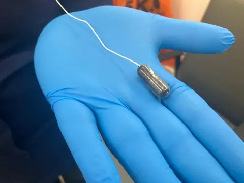 A hand in a blue rubber glove with a pill on the end of a string.