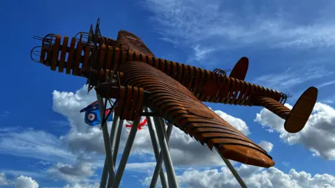 A metal structure made of oxidised steel - it's a life-size Lancaster bomber which is supported by grey tubes. The artwork appears to be banking and behind it is a blue sky with fluffy white clouds.