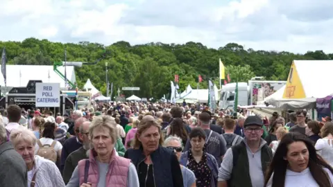Stuart Howells/BBC Large crowds walking through part of the Suffolk Show. The walkway is lined by stalls and marquees. There are trees in the background.