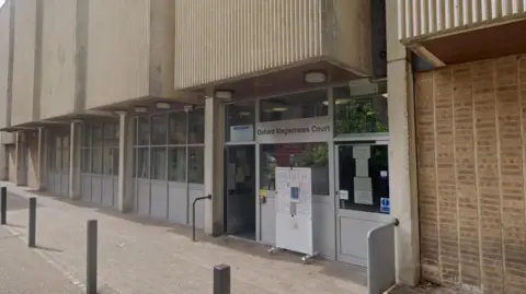 The front of Oxford Magistrates' Court. There are large glass doors under the brutalist concrete structure above the ground floor.