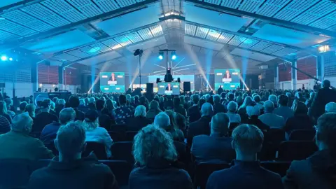 Paul Moseley/BBC A large crowd of people sat down bathed in turquoise light are watching a man address them from the stage. Large video screens are either side of the stage.