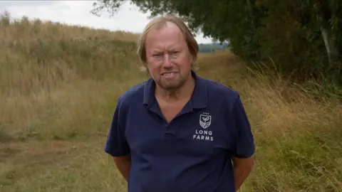 Jamie Niblock/BBC A man standing in a field on a cloudy day. He is smiling with his hands behind his back. He is wearing a blue polo shirt with the words 'Long Farms' printed on the chest. He has blond hair that has grown past his ears. 