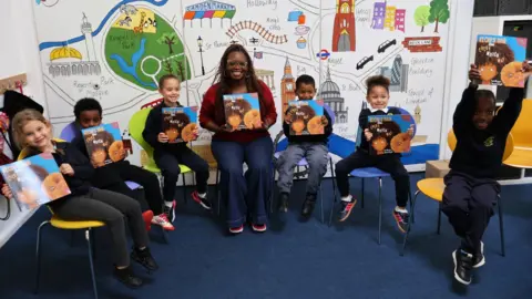 Bookmark The image shows a woman posing sat with 6 school-children, all in individual chairs, posing with the same book. They are smiling at the camera, and have a mural of London landmarks behind them.