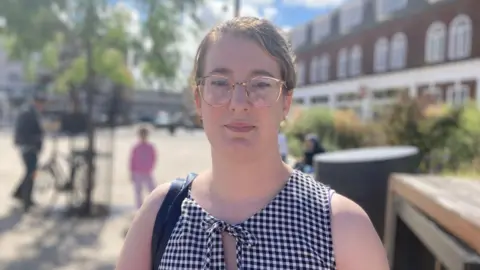 A young woman with her hair tied back, she wears glasses and a sleeveless black and white gingham blouse. 