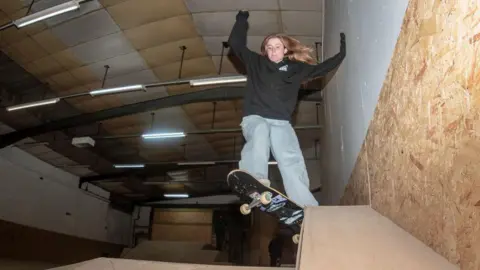 A woman riding a skateboard down a ramp. She is wearing a black jumper and blue jeans. There is a wooden skate park behind her.