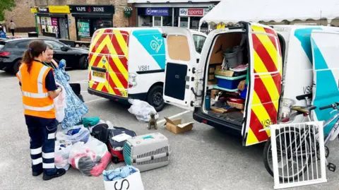 Two women in high-vis are stood beside large bags full of clothes. These are laid next to a van full of used items. There is a bike and a babygate leaning against the van. They are in a carpark in front of shops and a pub.