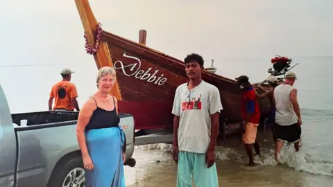 Margaret Garlick Margaret Garlick standing next to a Thai fisherman on the beach. Behind them is a boat with the name Debbie inscribed on it.