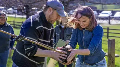 Yorkshire Dales National Park Authority A group of people in a grassy area planting a young tree, with one person holding the trunk while another supports the soil‑covered root ball, and others standing nearby against a backdrop of trees and a wooden fence on a sunny day.