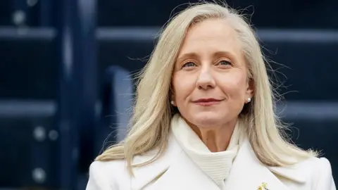 Abigail Spanberger is wearing a white coat and white scarf and pearl stud earrings. Her blonde hair is loose around her shoulders. 
