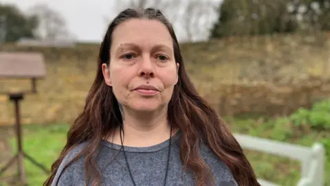Jo, a woman, with long brown hair, wearing a grey jumper, looking into the camera. She is standing in a garden with grass in the background.