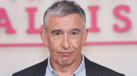 Steve Coogan with greying hair, clean shaven wearing a white Oxford-style shirt and a charcoal grey jacket