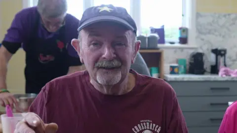 Dave Ackrell looks towards the camera wearing a burgundy tshirt and brown baseball hat. He has a grey moustache. He is holding his hand up towards the camera.