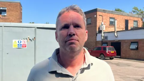Tony Fisher/BBC A man with stubble and short hair wears a polo top and is looking into the camera. Behind him there is grey hoarding on an industrial site. There is a red car in the background parked by a brick building with small chimneys coming out of the side.