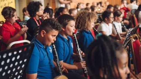 Soul Media Children from Horfield CE Primary school holding clarinets and one with a recorder, wearing school uniform and focussed on music in front of them. They look like they are mid-playing.