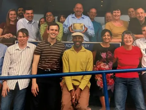 A number of people stood in front of the camera with a man in the middle holding an award.