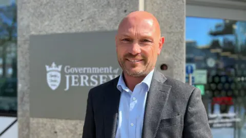 Ben Harvey stands outside a modern concrete building next to a large sign that reads “Government of Jersey” with the island’s crest. Glass windows reflect nearby trees and a white vehicle. Part of a post box graphic is visible on the left side of the frame. Bright daylight casts strong shadows on the scene.