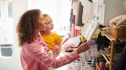 A woman holding a young girl in her left arm. They are both looking up at a central heating boiler. 