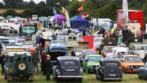 Boston Classic Car Club Rows of classic cars on a field. There is a Land Rover in the foreground, along with two vintage Volkswagen cars - one painted bright orange - the other bright green. There are a number of stalls to one side of the field.