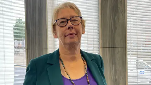 A women with short blonde hair in a green jacket and purple top. She is wearing black glasses and a beaded necklace. 