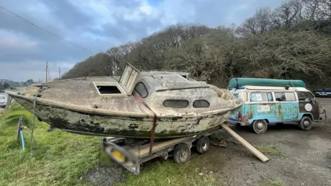 A yacht without a mast and covered in mould on a trailer with an old campervan.