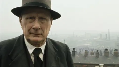 Getty Images Artist LS Lowry standing in front of his terraced house home in Pendlebury, with the chimneys and factories of Salford in the background