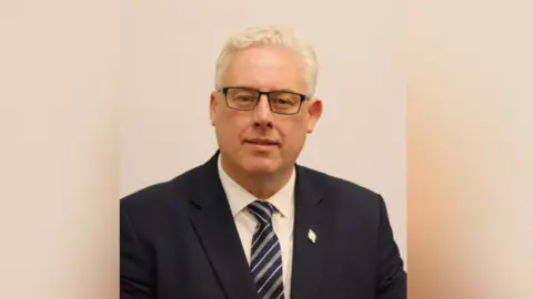 A man with white hair and dark rimmed glasses is looking into the camera. He is wearing a dark blue formal blazer, a white shirt and a striped tie.