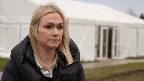 Jo Lowry has shoulder-length blonde hair and is wearing a black hooded puffer coat. She is standing in front of a large white marquee tent which has one of its glass double-doors propped open. 