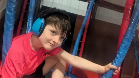 Sarah Duncan A young boy wears a red top and black shorts. He has blue earmuffs on. He sits in a soft play area, holding on to a blue foam pole with his left hand. 