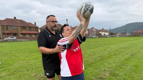 Carl guides his son, Jak, for the line-out throw as Jak lifts the ball. Cal is wearing a black shorts and t-shirt combo while Jak has a red and white Lions top on and blue shorts. They are stood on a grassy pitch and there are houses behind them.