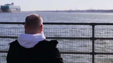 Phill is pictured from behind, sitting and looking out to sea, with a boat in the background. He has short fair hear and is wearing a white hoodie and a black coat.
