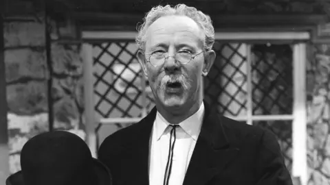 A black-and-white photo of a man pictured from the chest up. He is in his 50s, but made-up to look older, with white hair and a drooping white moustache. He is wearing a black jacket and a white shirt, glasses, and is holding a black bowler hat in his right hand, visible at the bottom of frame as he holds it up in front of him. He is standing in front of a stone-walled house with a leaded, diamond-pattern window, with a framed painting visible on the wall through the window, and he is smiling at the camera.