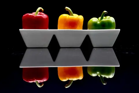 Mick Bracken Three peppers in a white bowl reflected on a shiny surface, pictured against a black background