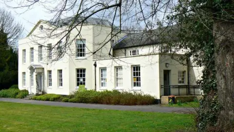 Brian Robert Marshall/Geograph The outside of a large cream coloured building, with several windows. It has a grey roof, and a tree to the right, with branches at the front of the image. There is grass in front of the building. 