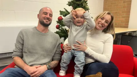 A man and a woman on a red sofa. Between them is a toddler. He is smiling and holding his hands up to his head. 
