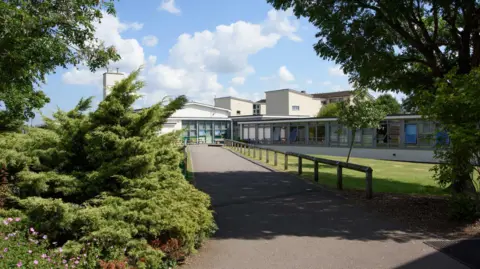 Exterior shot of Wellsway School in Keynsham. The pathway leading to an entrance to the school. There are trees either side and a lawn to the right hand side of the path.
