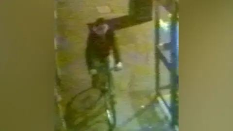 A grainy still from CCTV which shows a man wearing a dark jacket cycling on the pavement past a phone box