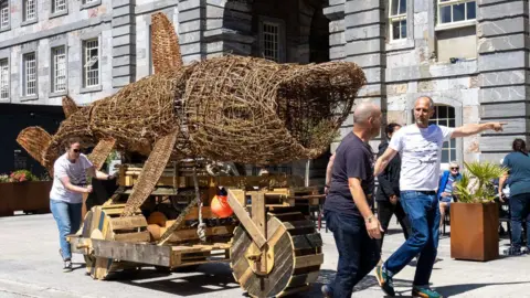The wicker shark is being transported by a wooden trailer with wooden wheels. It is being guided at the front by two men and a woman is steering from the tail end area. They are pushing the model past old brick buildings. The model shark has its large mouth open as if it is feeding.