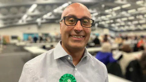 Elizabeth Baines/BBC A man with glasses wears a grey shirt. Behind him election counts are taking place.