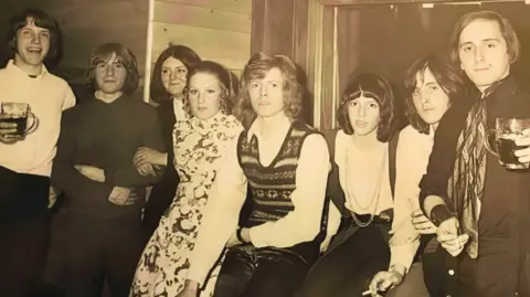 David Maynard David Bowie, who has curly dark blonde hair, poses for a photo backstage at a club surrounded by seven teenagers including four boys and three girls. 