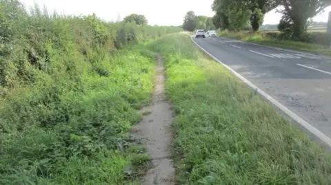 The current path. It is narrow and has overgrown vegetation encroaching on to it. There are several cars in view on the main road, which is flanked by trees on the other side