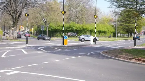 A zebra crossing over a wide road with four black and white striped posts visible with yellow lights at the top. It is a bright day with blue sky. Cars can be seen on the road turning away from the crossing.